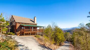 A log cabin with a porch and a chimney, surrounded by trees and a clear sky.