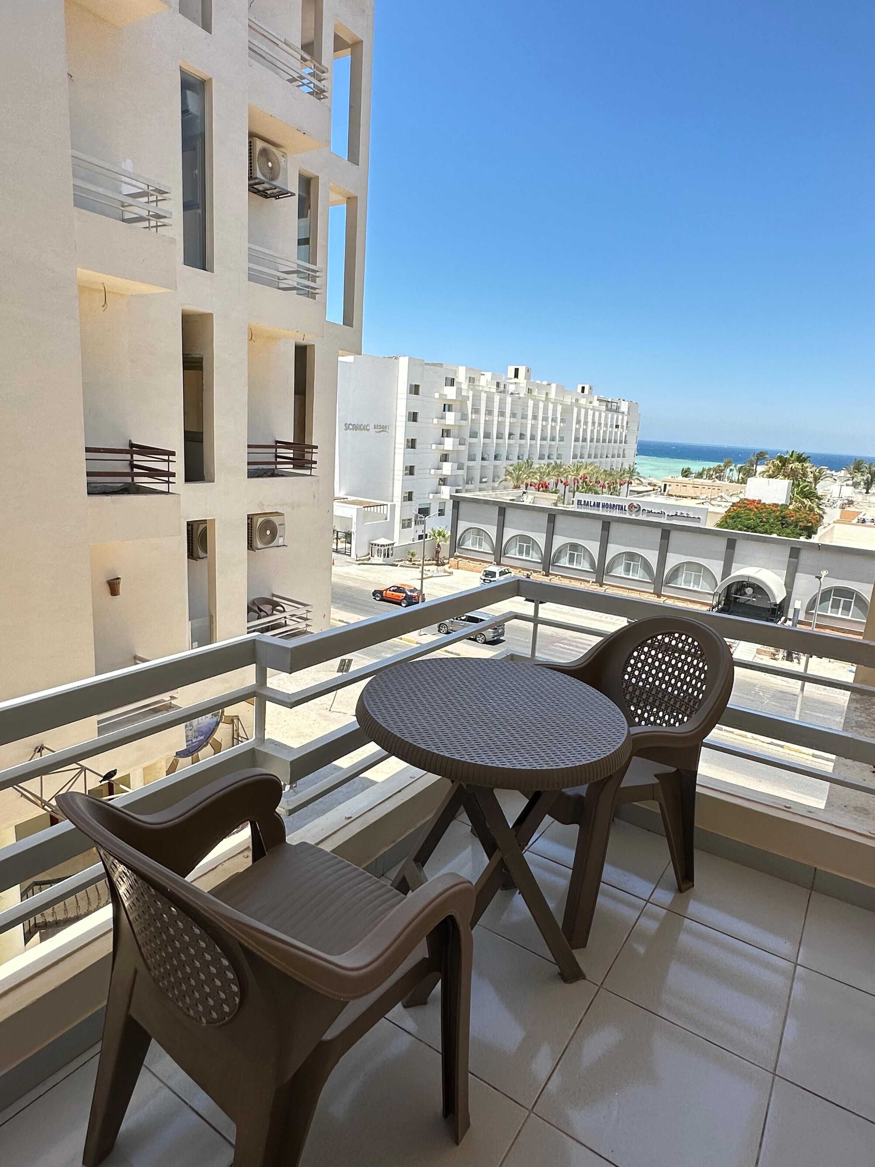 A balcony with two chairs and a table, overlooking a beach and a hospital building.