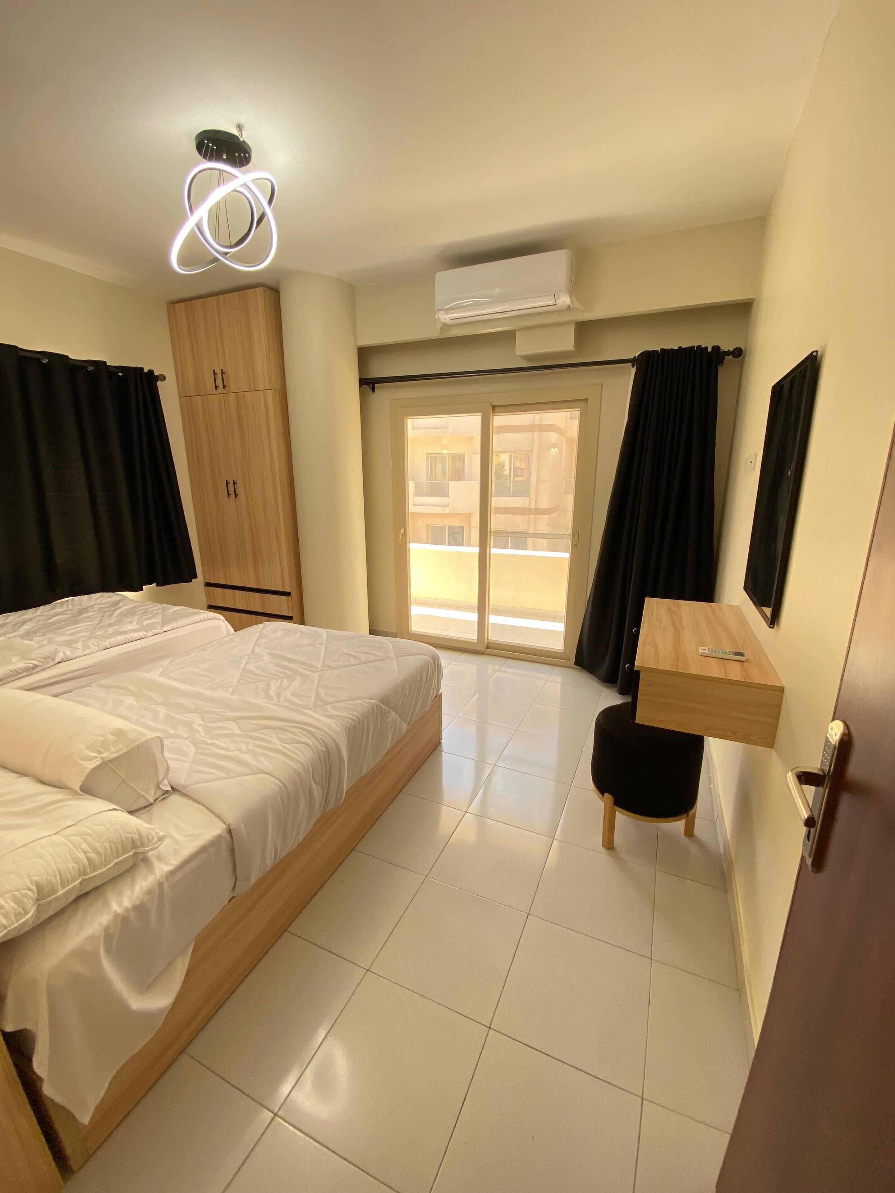 A bedroom with a bed, a wooden wardrobe, a desk with a chair, and a sliding glass door to a balcony.