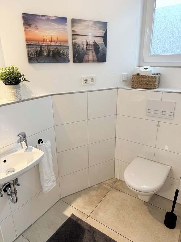 A modern bathroom with a sink, toilet, and a small window. There are two wall-mounted paintings and a potted plant on the countertop.