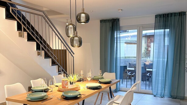 Un comedor moderno con una mesa de madera dispuesta para cuatro personas, sillas blancas y una escalera con barandilla de metal.