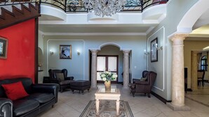 Ein geräumiges Wohnzimmer mit hohen Decken, einer Kristallleuchtre und einer roten Akzentwand. Es gibt zwei Ledersofas, einen Couchtisch und eine dekorative Vase.