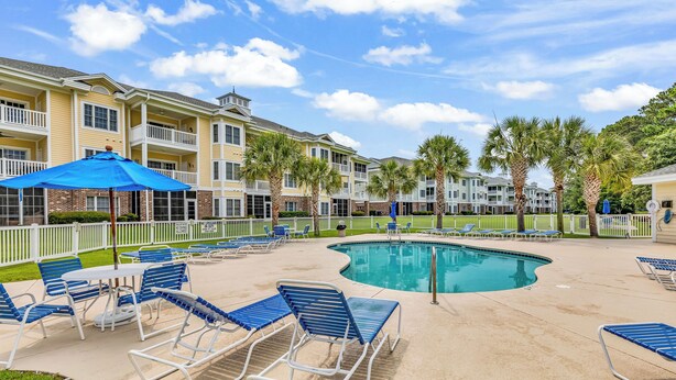 A pool area with lounge chairs and umbrellas, surrounded by a white fence and palm trees. - Myrtlewood 2-bedroom condo.  Pool in the backyard (Myrtle Beach)