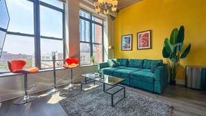 Un salón moderno con un sofá verde, dos sillones rojos, una mesa de centro de vidrio y un ventanal con vistas a la ciudad.