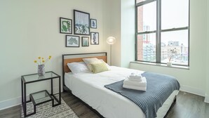 Un dormitorio moderno con un ventanal grande, una cama con ropa de cama blanca, una mesita de noche con un jarrón de flores y cuadros en la pared.