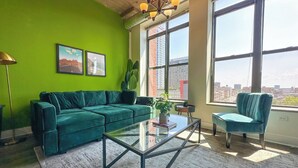 Una sala de estar moderna con un sofá verde, una mesa de centro de vidrio y vistas de la ciudad a través de grandes ventanales.