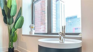 Un baño moderno con un lavamanos redondo, una planta en maceta y vistas a un paisaje urbano a través de un ventanal grande.