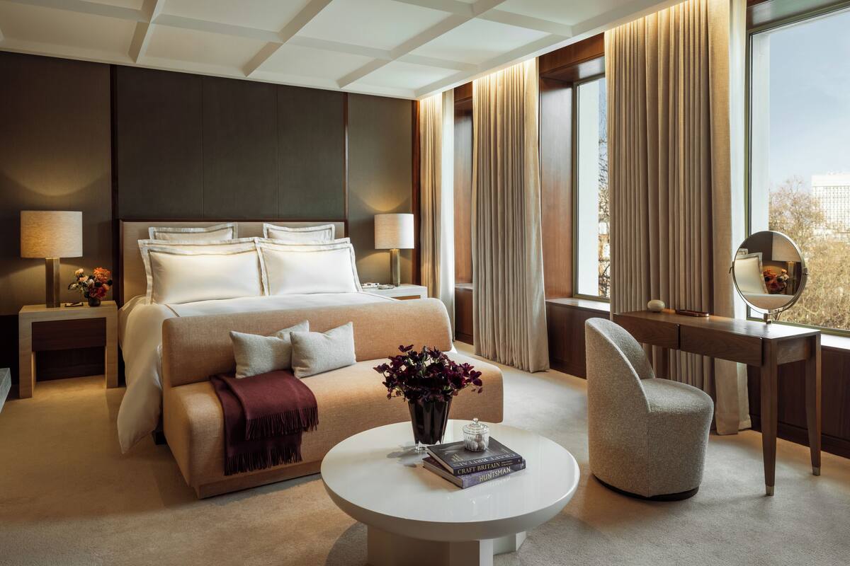 A modern hotel room with a large bed, a sitting area with a coffee table, a desk with a mirror, and a view of the city through large windows.