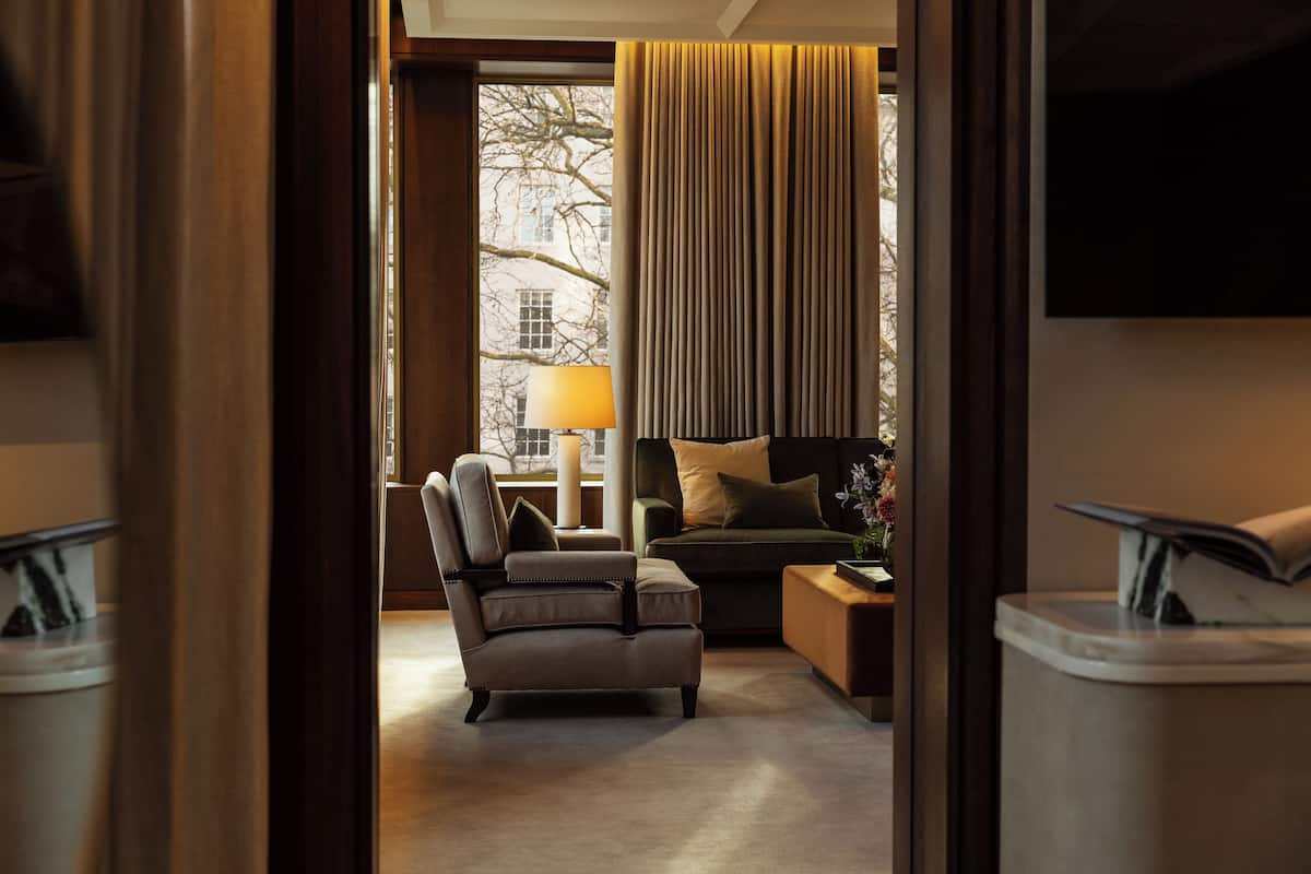 A hotel room with a leather armchair, a sofa, a side table with a lamp, and a view of the outdoors through a window with curtains.