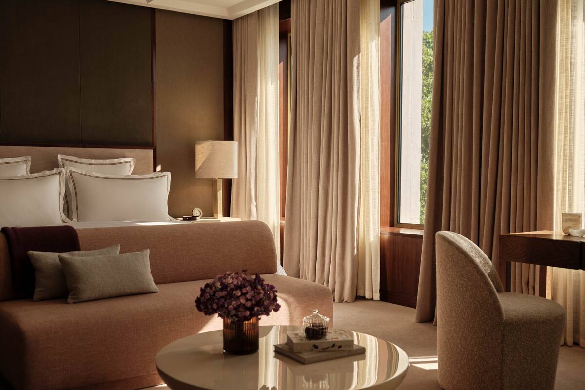 A hotel room with a bed, a sofa, a coffee table, and a chair. There is a window with curtains, a lamp, and a vase with flowers on the coffee table.