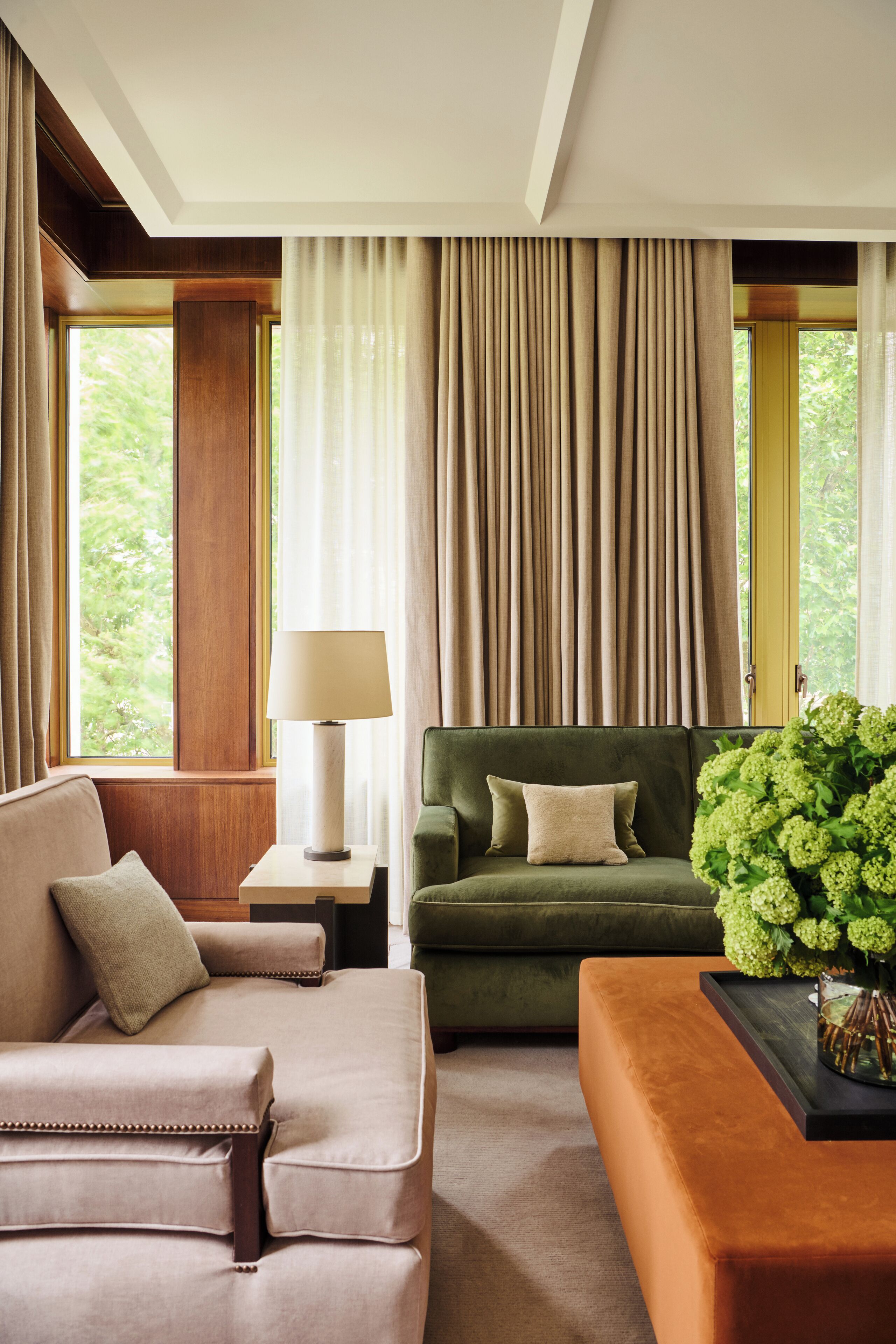 A hotel room with a green sofa, a beige armchair, a small table with a lamp, and a large window with curtains.