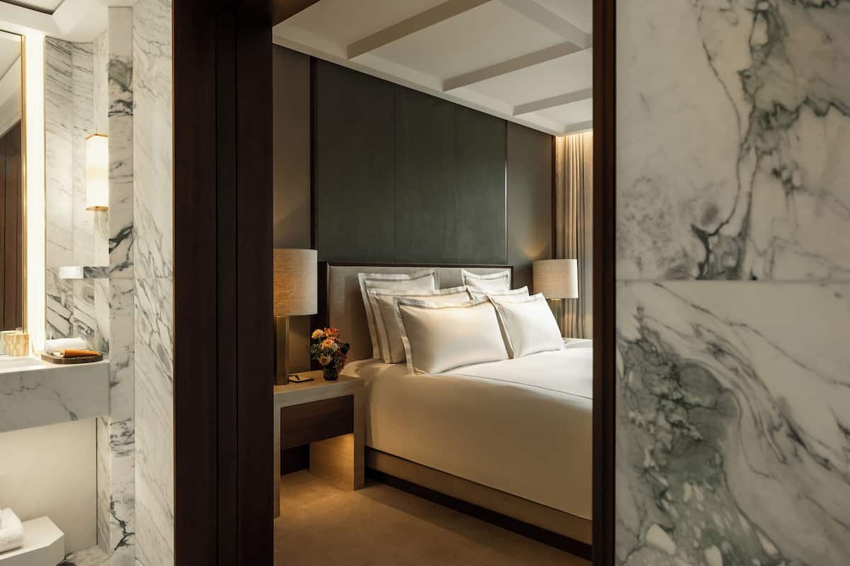 A hotel room with a marble wall, a bed with white linens, a nightstand with a lamp, and a wooden headboard.