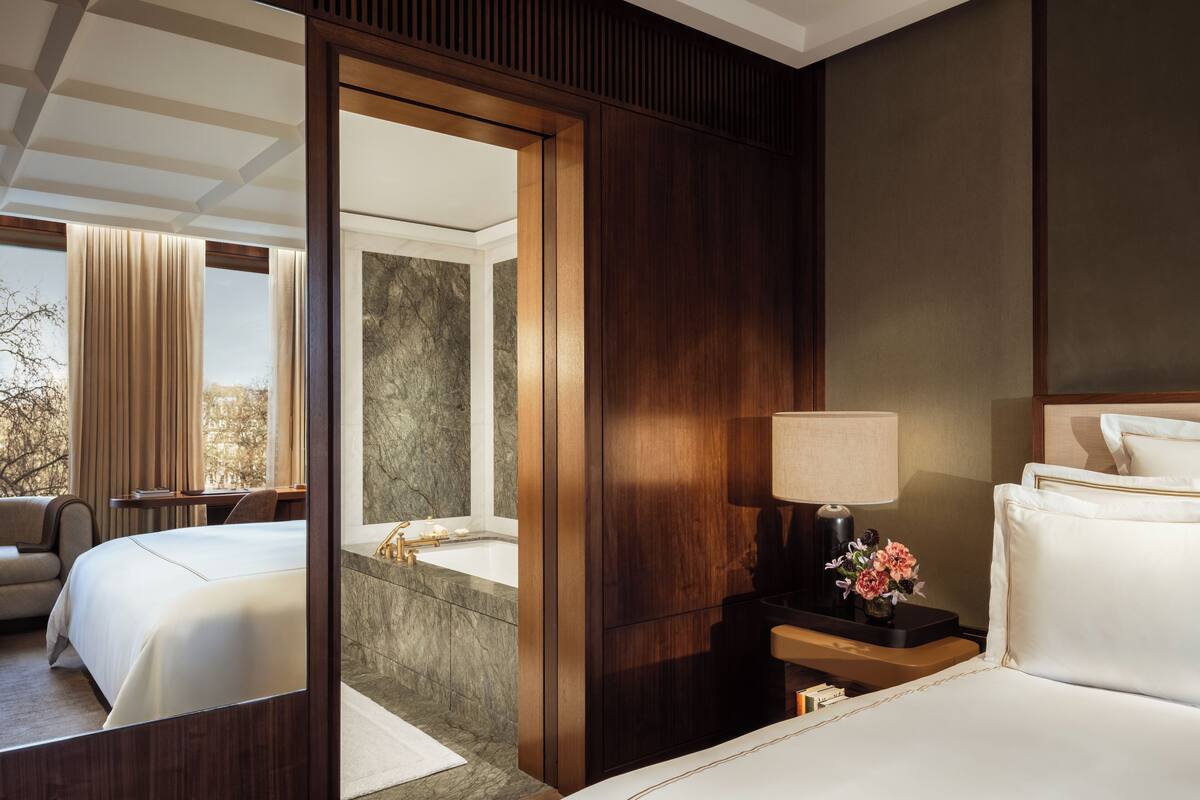 A hotel room with a large bed, a bedside table with a lamp, a mirror, and a view of another room with a bathtub and a window.