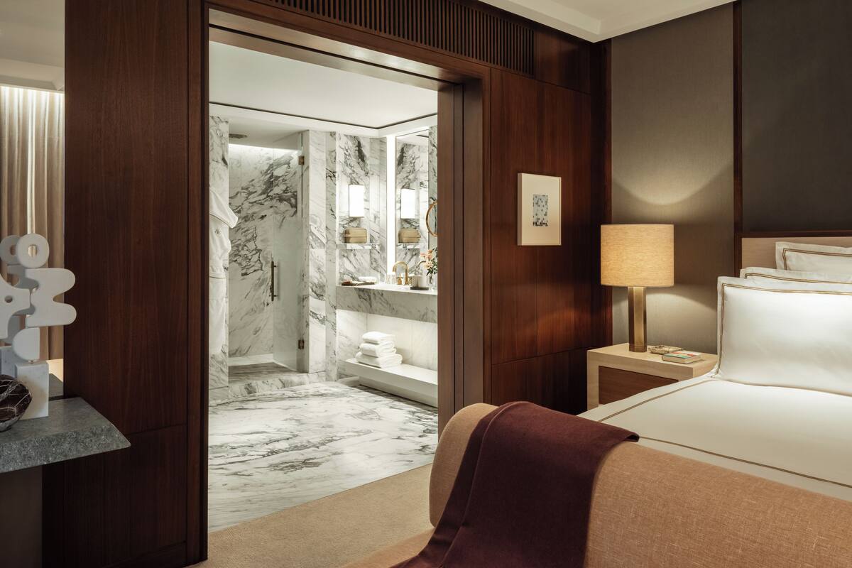 A hotel room with a marble bathroom, a bed with a headboard, a bedside table with a lamp, and a wooden wardrobe.
