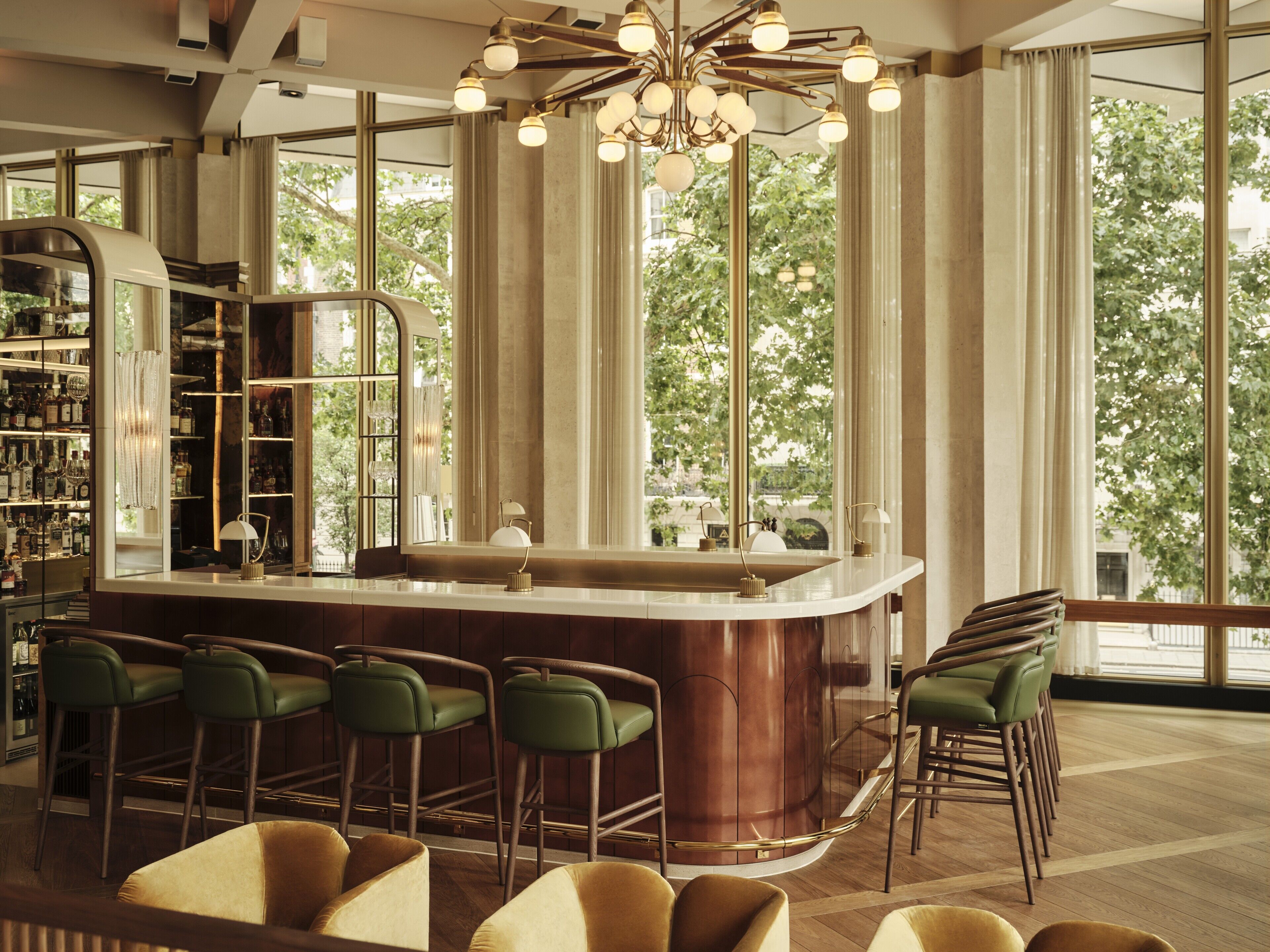 Een bar met een koperen toog, groene barkrukken en een groot raam met uitzicht op bomen.