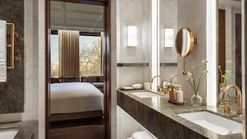 A modern bathroom with a large mirror, two sinks, and a bed visible through an open door.