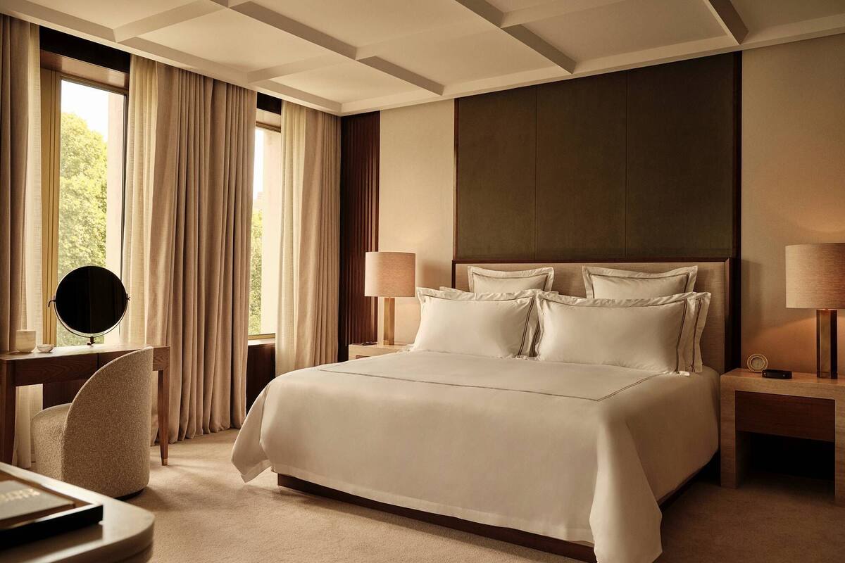 A hotel room with a large bed, two bedside tables, a desk with a chair, a mirror, and a window with curtains.