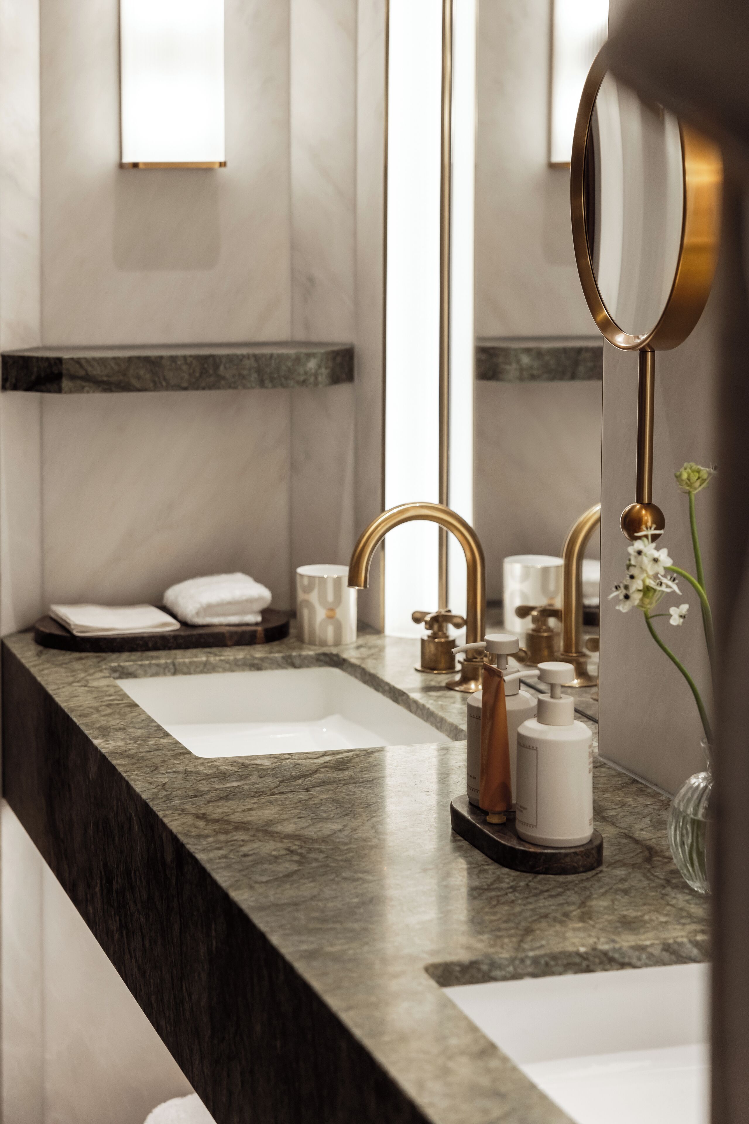 A modern bathroom with a marble countertop, two sinks, gold faucets, and a large mirror.