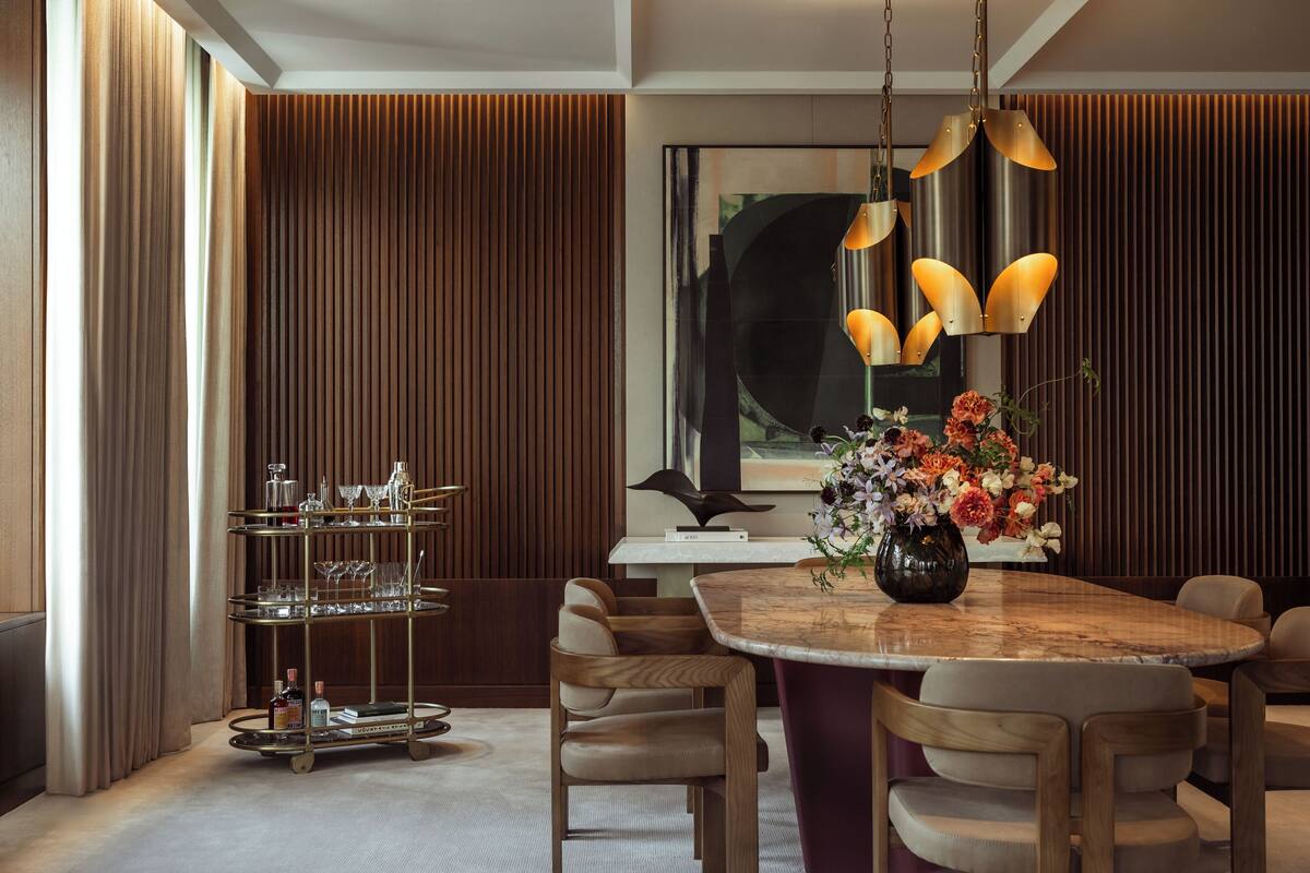 A modern dining area with a wooden table, multiple chairs, a glassware cart, and a floral centerpiece.