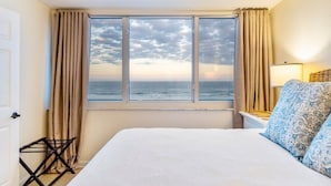 A hotel room with a bed, a window with a view of the sea, a lamp, and a small table.
