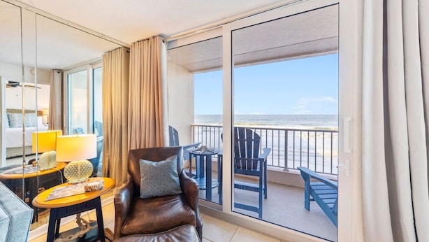 A balcony with a view of the beach, a leather chair, a small table with a lamp, and a sofa.