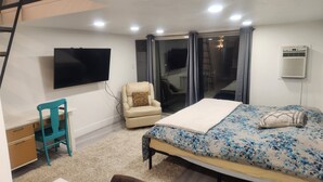 A bedroom with a bed, a desk, a chair, a TV, and a sliding glass door to the outside.