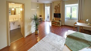En rummelig stue med trægulv, et stort vindue med gardiner, et fladskærms-tv, et skrivebord og en potteplante.