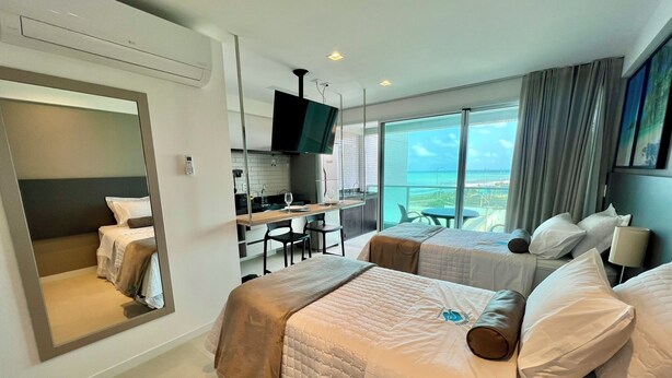 Een hotelkamer met twee bedden, een kitchenette, een televisie en een balkon met uitzicht op zee.