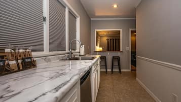 A kitchen with a marble countertop, a sink, and a window with blinds. There are two stools and a mirror above the sink.