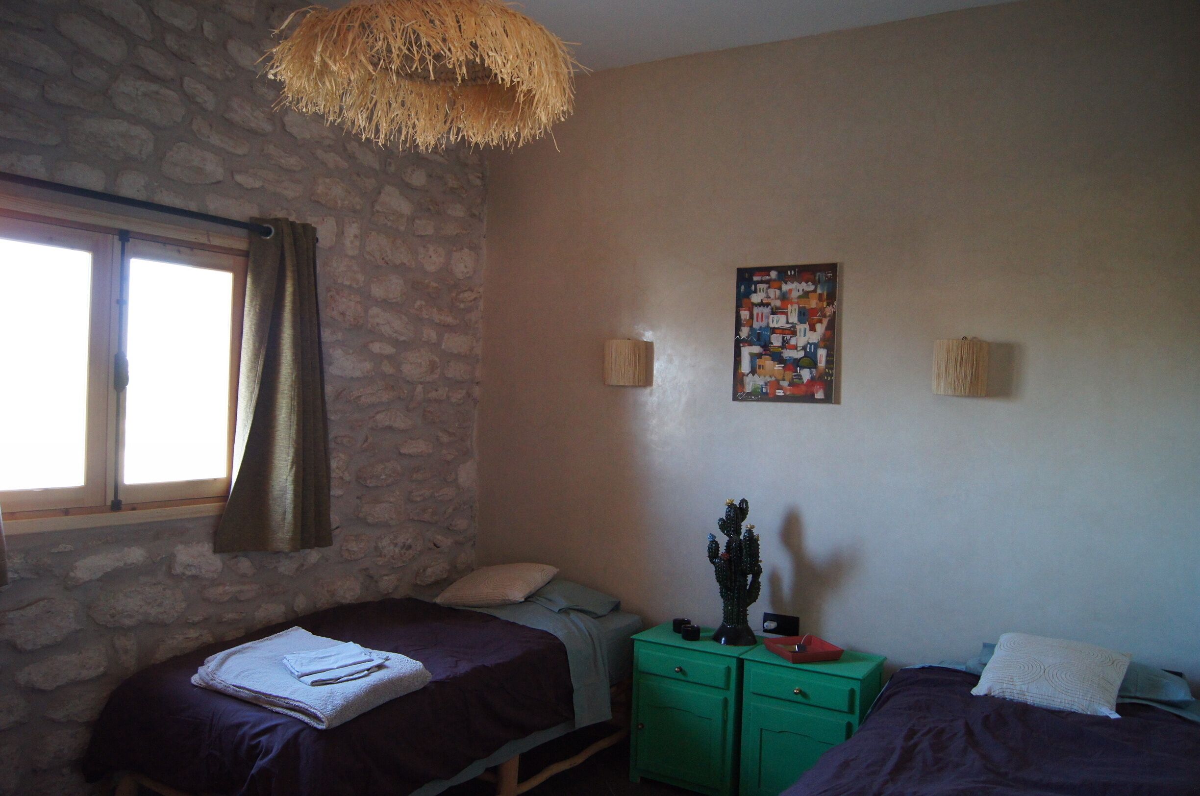 A bedroom with two beds, a stone wall, a window with curtains, a green nightstand, and a wall-mounted lamp.