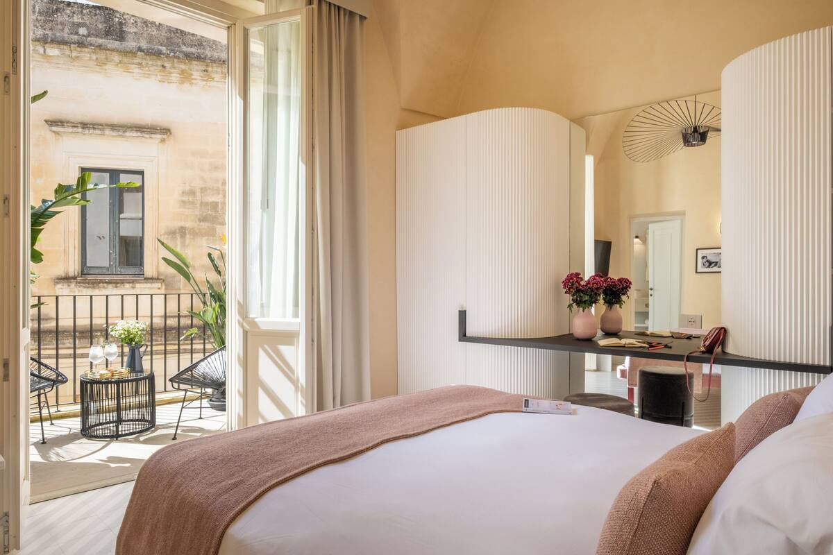 Une chambre d’hôtel avec un lit, un bureau doté d’un miroir, un petit balcon avec des chaises et une vue sur le bâtiment extérieur.