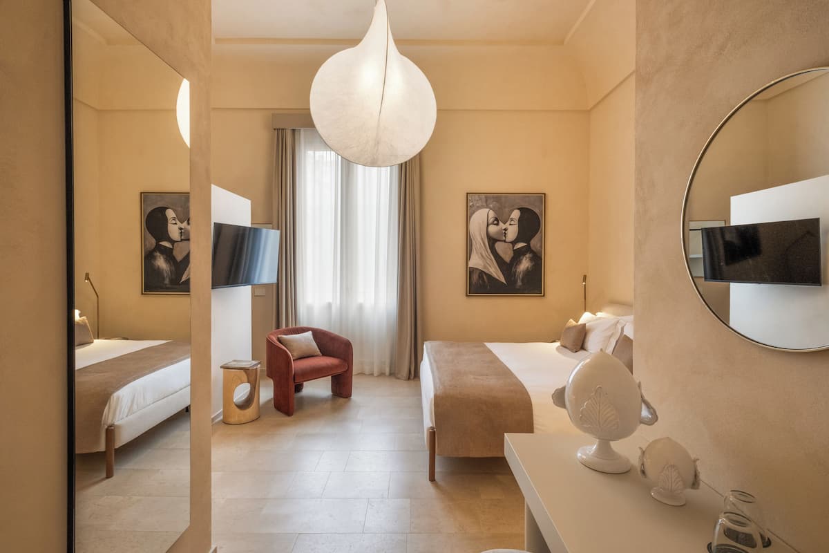 Une chambre d’hôtel avec deux lits, un coin salon avec un fauteuil rouge, un grand miroir et des œuvres d’art accrochées aux murs.
