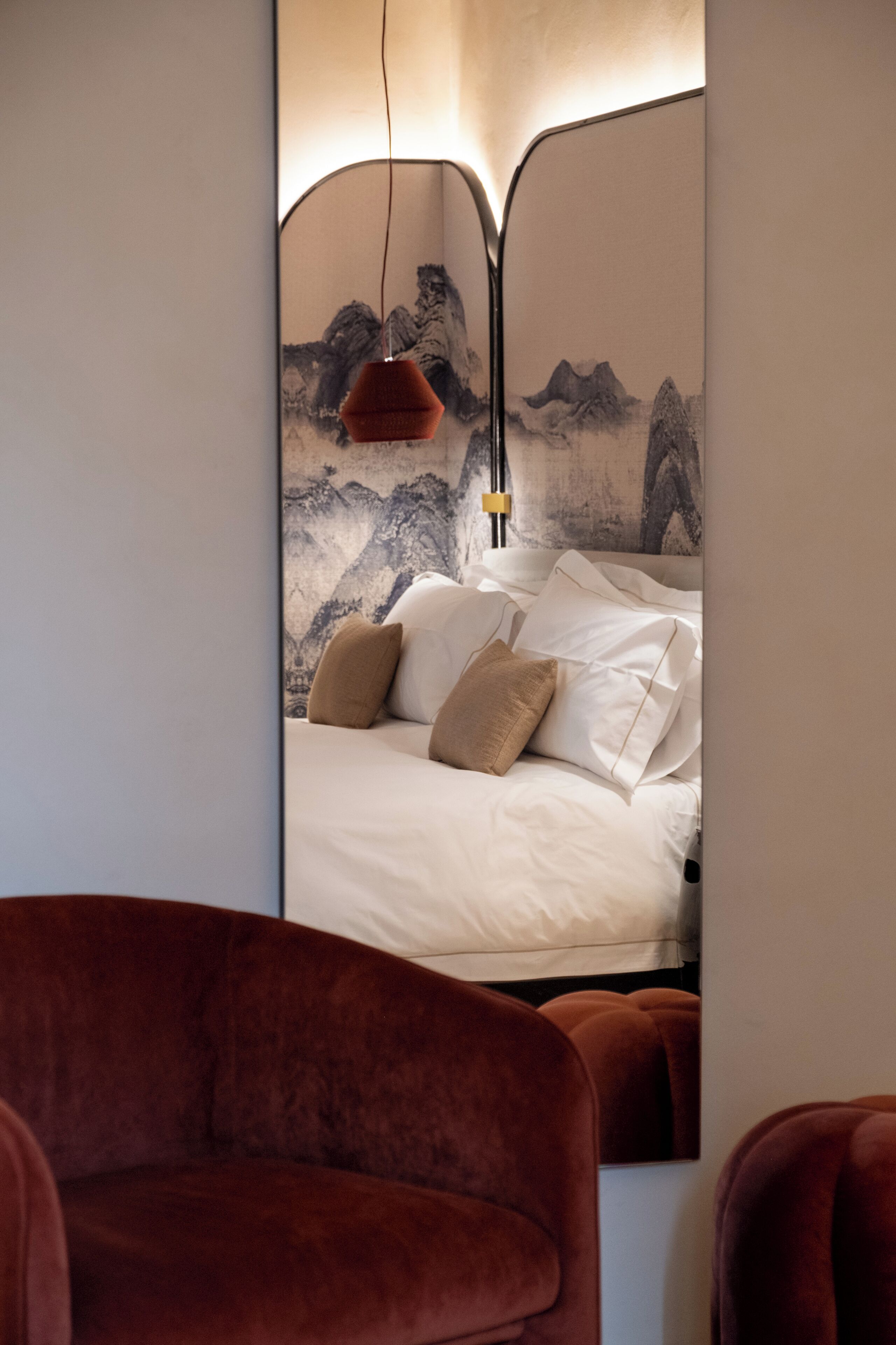 A bedroom with a bed, pillows, a red hanging lamp, and a wall with a mountainous landscape painting.