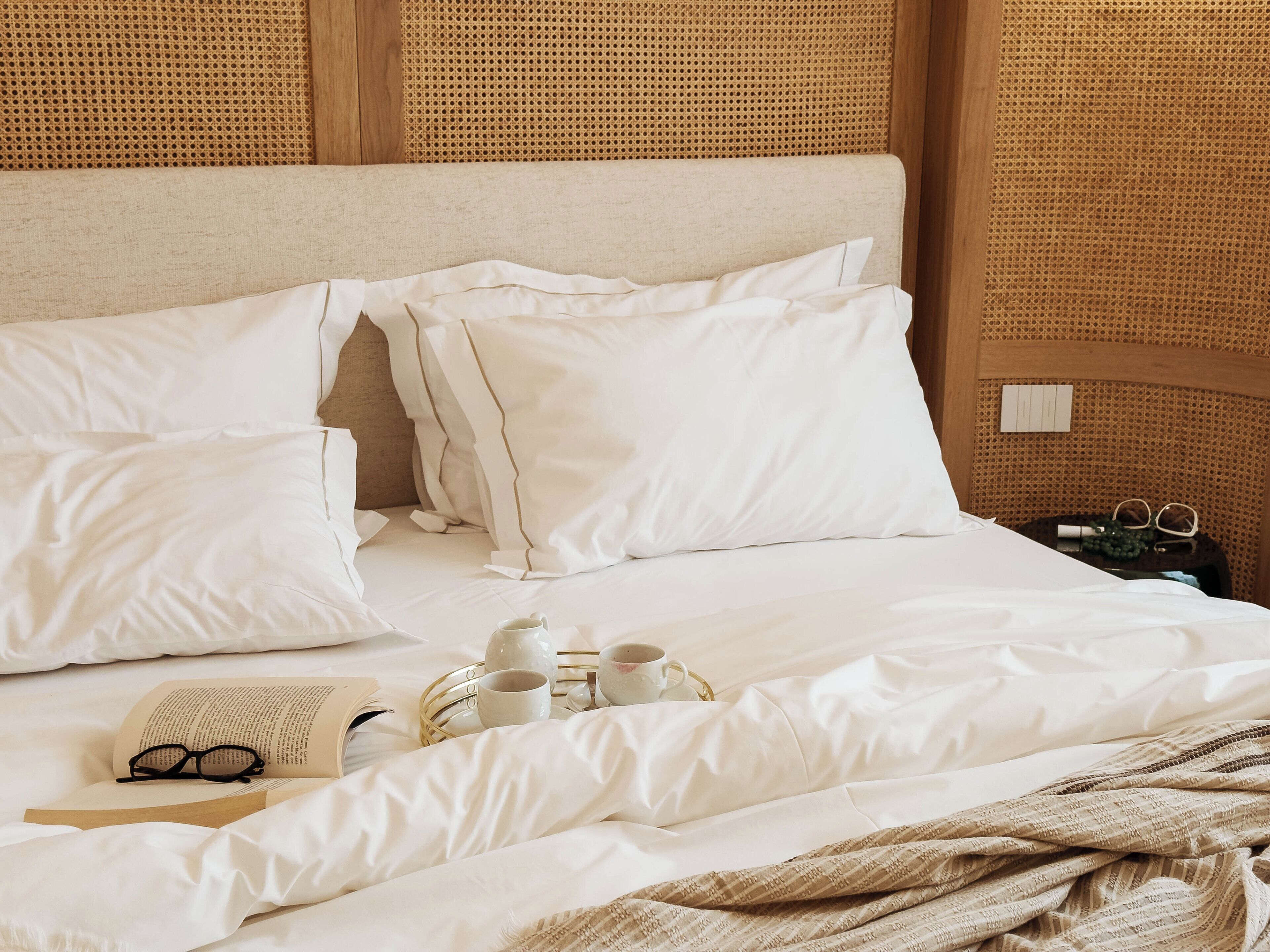 Cama bem arrumada com lençóis brancos, mesa de cabeceira com uma bandeja contendo xícaras e um livro, e cabeceira de tecido.
