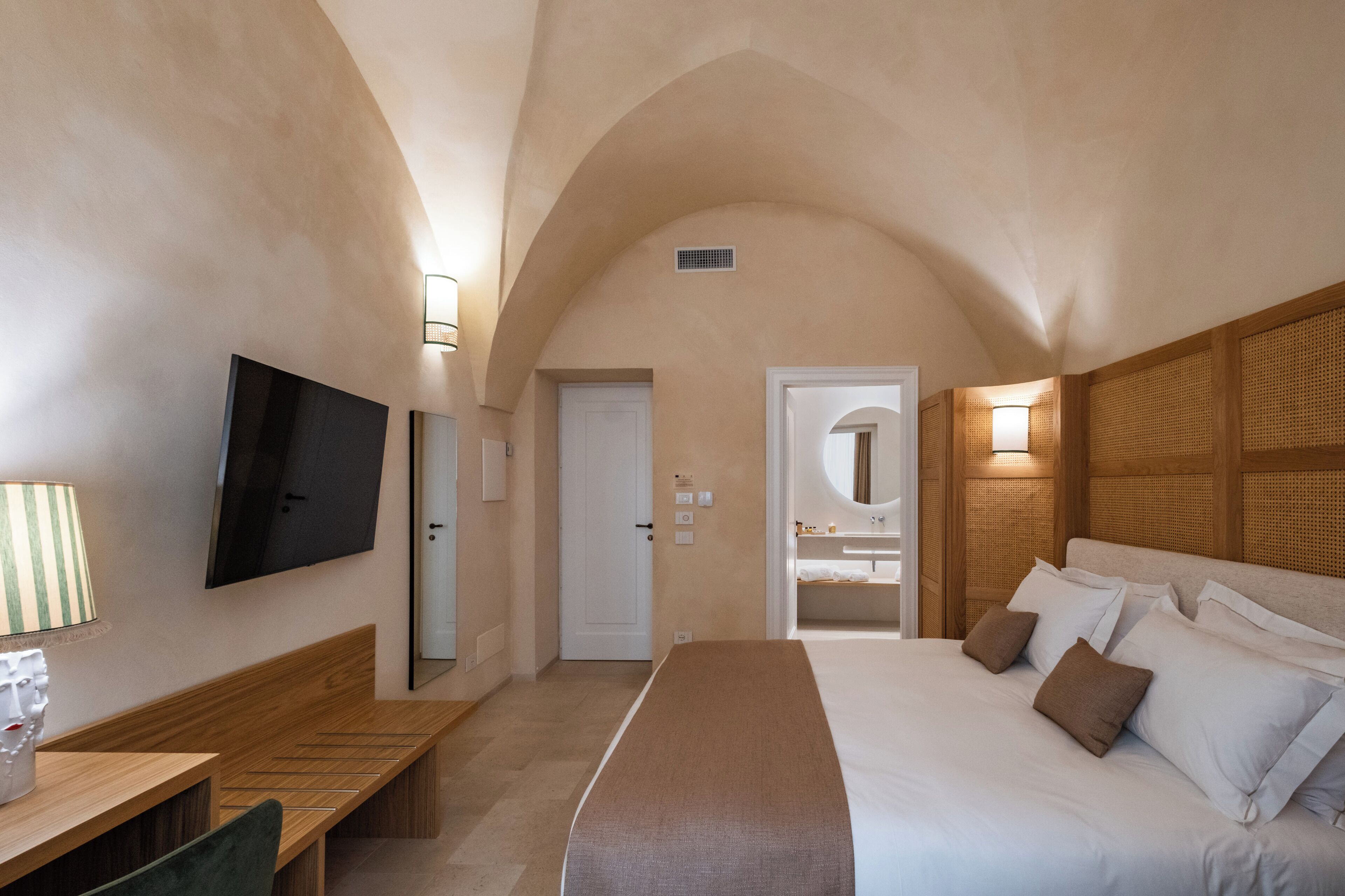 A bedroom with a large bed, a TV mounted on the wall, a wooden bench, and a door leading to a bathroom.