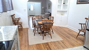 Een eetgedeelte met een houten tafel en stoelen, een keuken met een fornuis, en een witte kast met glazen deuren.