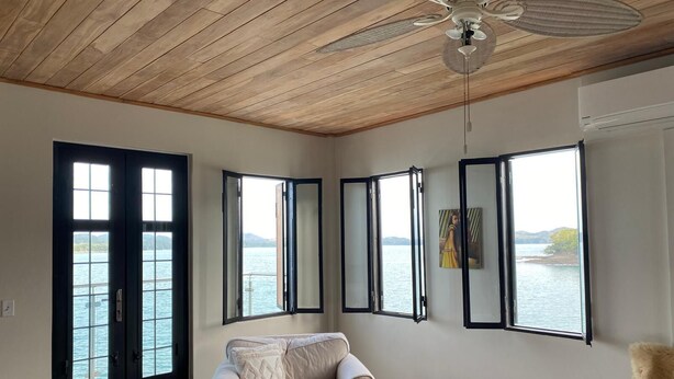 A room with a wooden ceiling, a beige armchair, a table with a book, and large windows with black frames overlooking a body of water. - Charming 2-bedroom, 2.5 bath Over-water House on Secluded Beach! (Boca Chica)