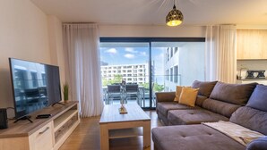 A modern living room with a flat-screen TV, wooden coffee table, and a sofa with cushions. A sliding glass door leads to an outdoor balcony with a view of buildings and greenery. - Conchas do Funchal by Madeira Real (Funchal)