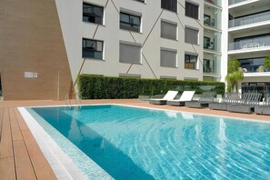 Une piscine avec des chaises longues, une terrasse en bois et un bâtiment moderne doté de balcons en arrière-plan.