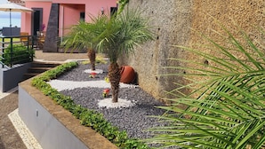 Un bâtiment rose avec un toit en tuiles, entouré d’un jardin aménagé avec des palmiers et un mur de pierres.