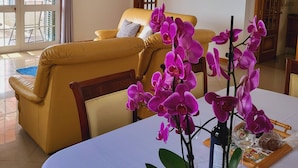 Un salon avec un canapé jaune, une table à manger recouverte d’une nappe blanche et des orchidées violettes.