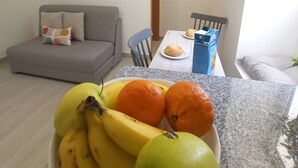 A living room with a grey sofa, a painting of a car on a beach, a dining area with a table and chairs, and a bowl of fruit on the counter. - Studio Theo Vista Montanha (Calheta (Madeira))