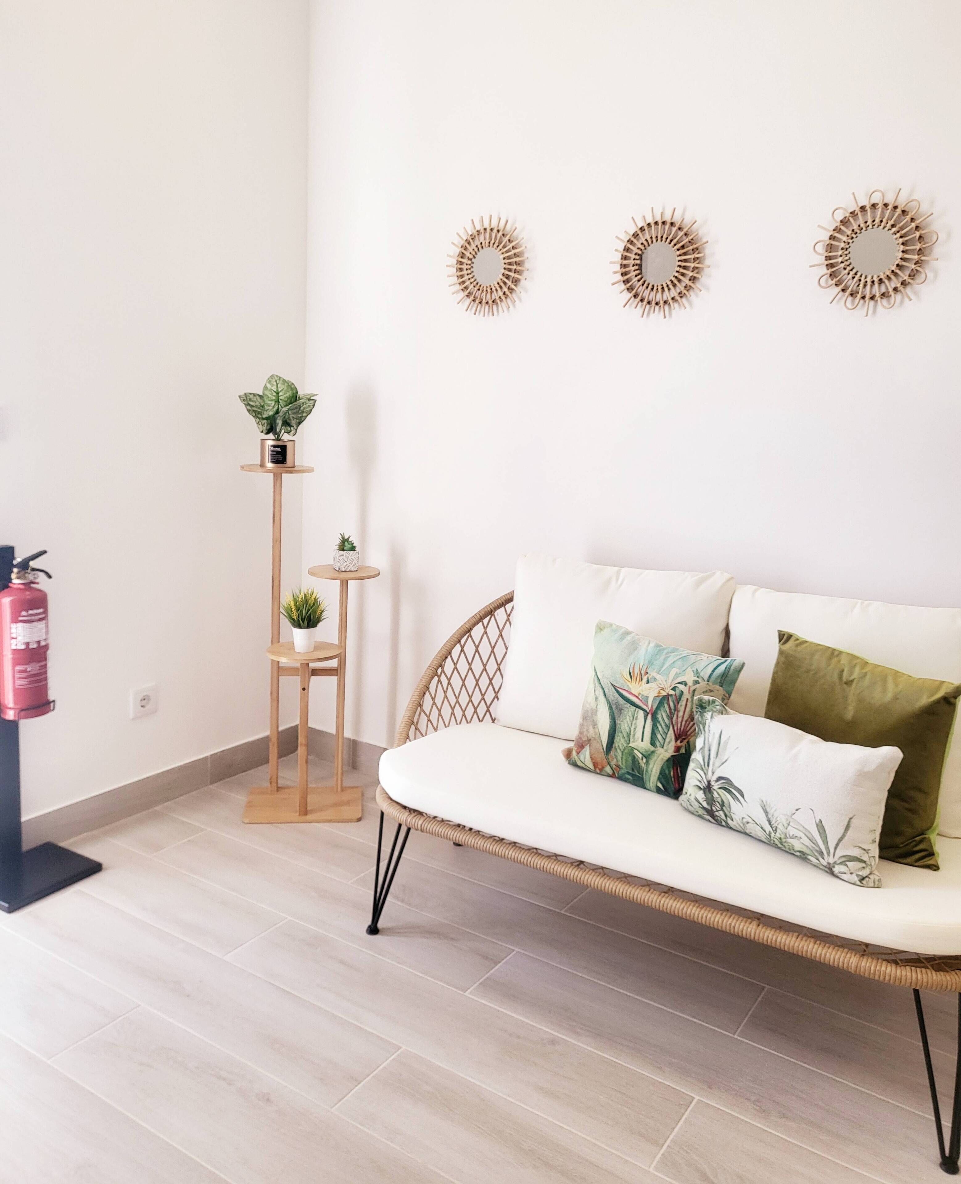 Una sala de estar moderna con un sofá de mimbre, dos espejos montados en la pared, una pequeña mesita de madera con una planta en maceta y un extintor de incendios en el suelo.