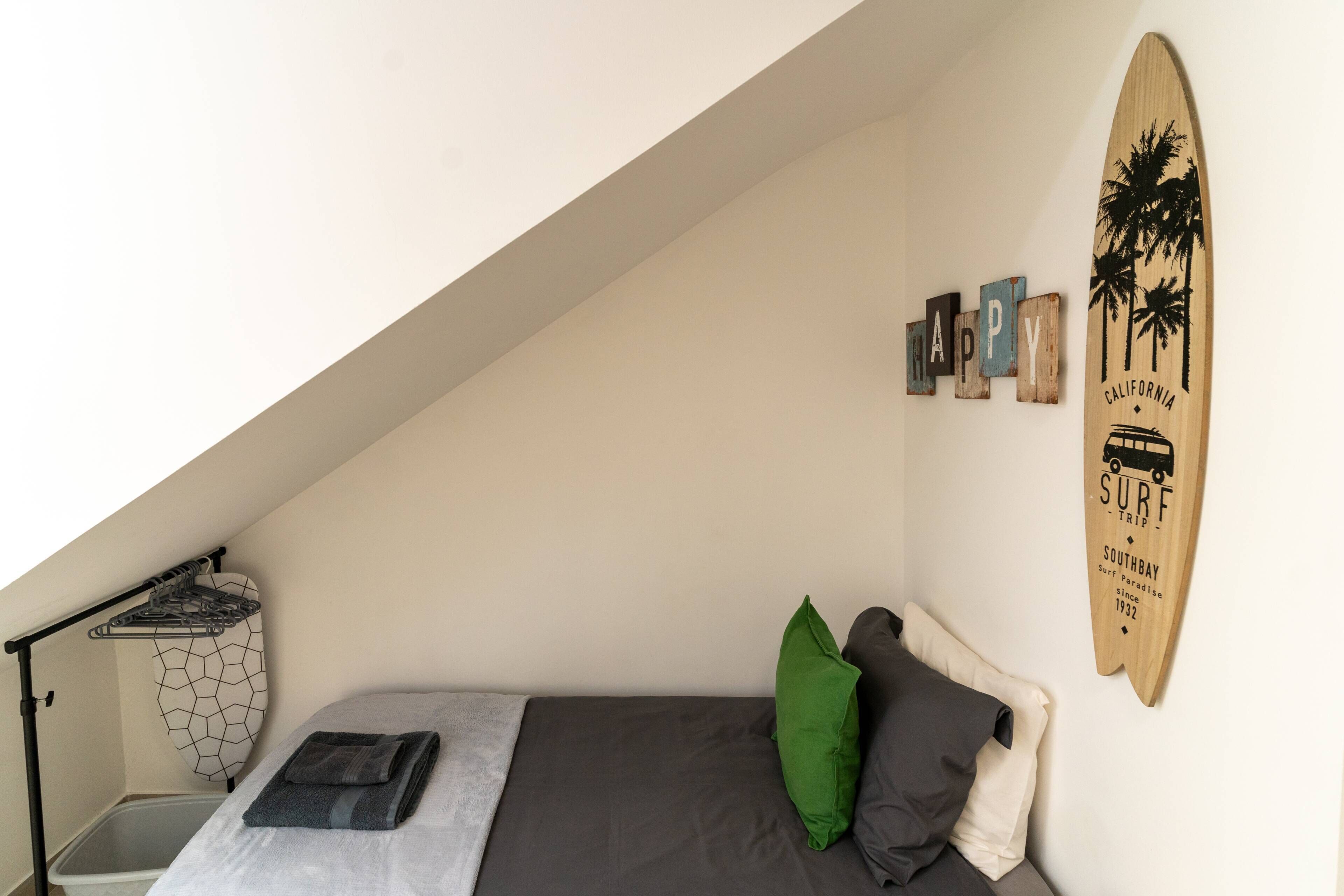 A small, cozy bedroom with a bed, pillows, a surfboard decoration, and a wall-mounted drying rack.