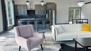 A modern kitchen with grey cabinets, a black countertop, and stainless steel appliances. There is a white sofa with a yellow pillow, a pink armchair, and a black leather ottoman.