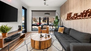 A modern living room with a round wooden coffee table, a sofa, a TV mounted on the wall, and a large window with a view of the street.
