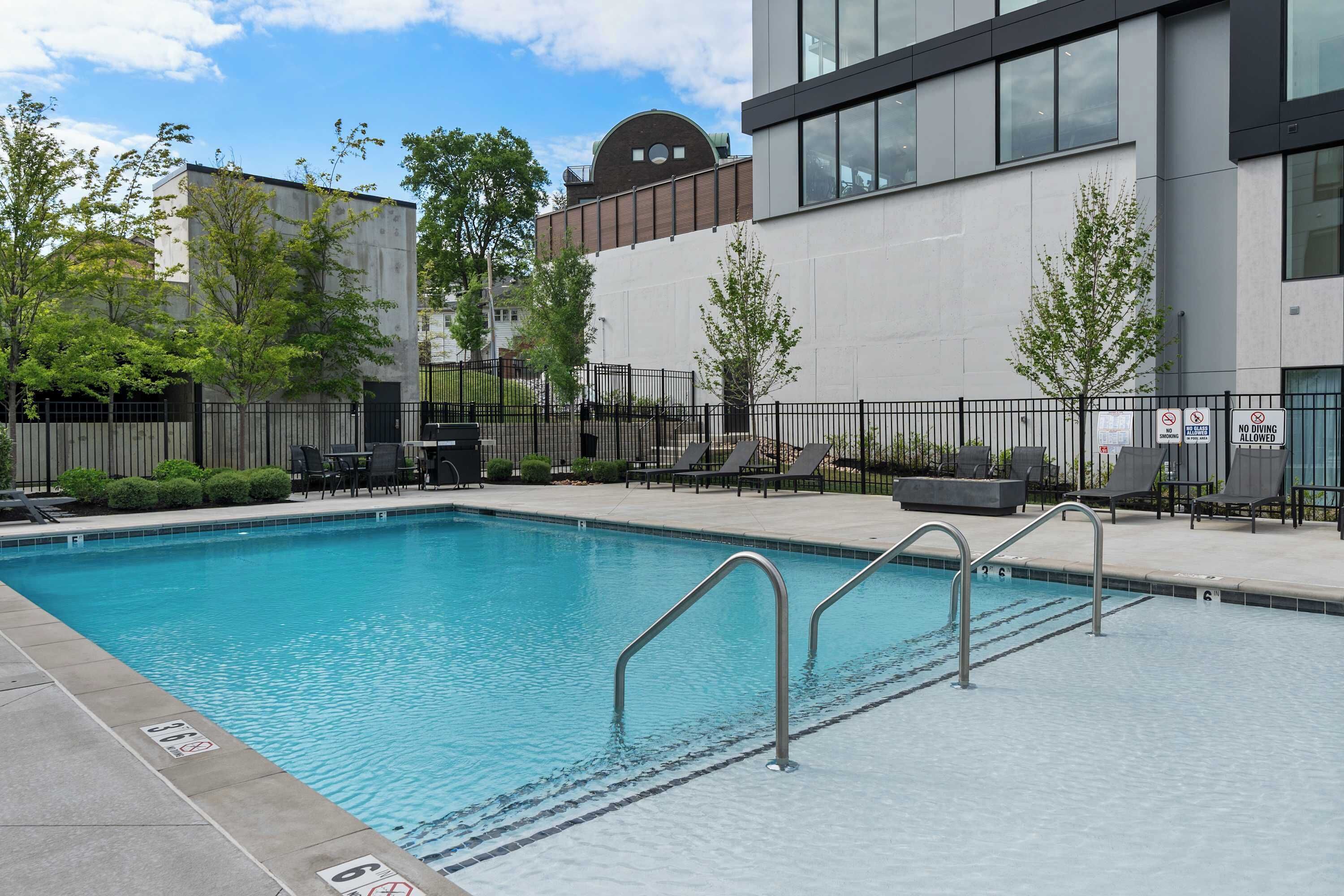 A rectangular swimming pool with metal railings, surrounded by lounge chairs and a fence.