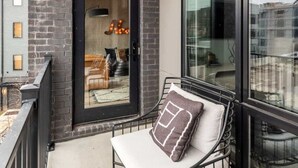 A balcony with a metal bench, a glass door leading to an interior space, and a view of the cityscape.