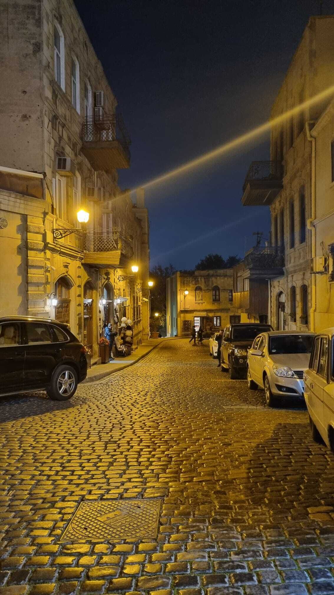 A cobblestone street at night with buildings on either side, street lamps, and parked cars.