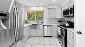 A modern kitchen with stainless steel appliances, white cabinetry, and a large window with a view of greenery. - Pet Friendly - Q2 & Domain - King Bed + Pool Table (Austin)